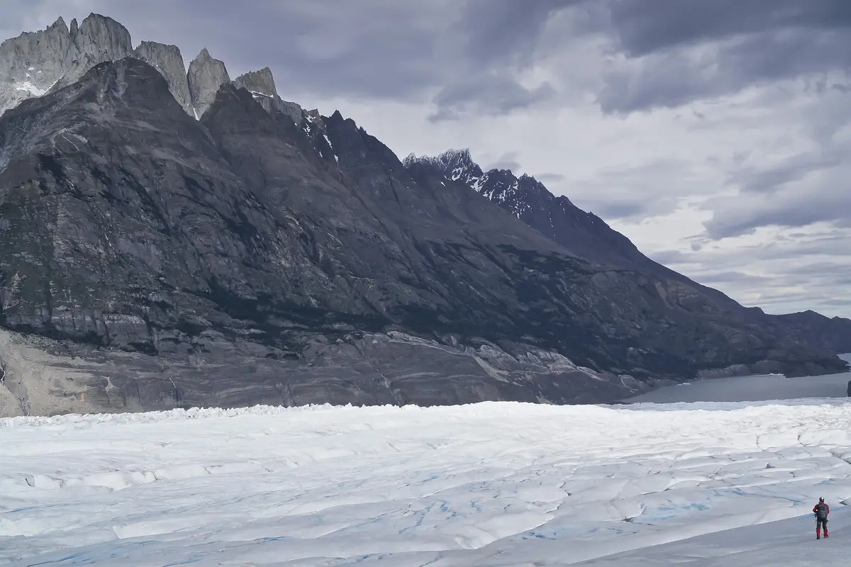 Un escursionista in rosso sulla superficie bianca e crepata del Ghiacciaio Grey, con scure montagne di granito che si ergono sul lato opposto sotto un cielo pesante