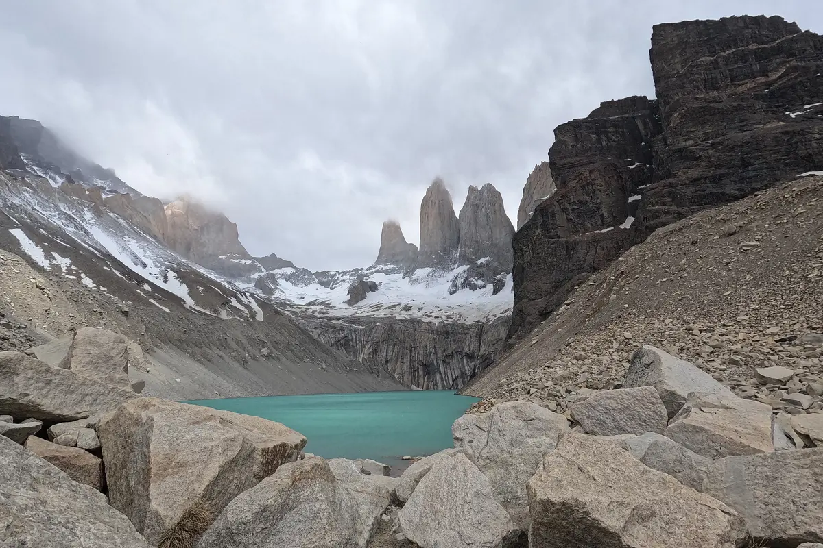 The three granite towers of Torres del Paine above a turquoise lake, seen across a boulder field with cloud on the peaks